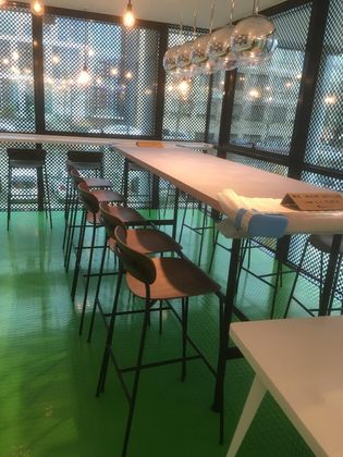 Une table haute et des chaises sont disposées sur un sol vert vif devant un mur vitré orné d'un grillage métallique et de suspensions lumineuses.