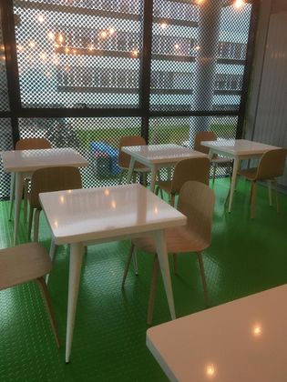 Une pièce lumineuse avec des tables carrées blanches et des chaises en bois clair disposées sur un sol vert texturé, devant de grandes fenêtres.