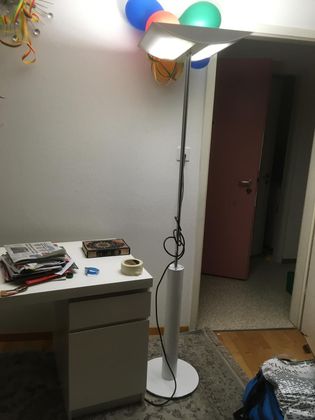 Un grand lampadaire moderne blanc, doté de deux panneaux rectangulaires lumineux, se dresse à côté d'un bureau blanc et d'une porte.