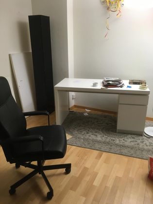 Une chaise de bureau noire fait face à un bureau blanc, avec un tapis gris, un parquet et une grande enceinte noire sur la gauche.
