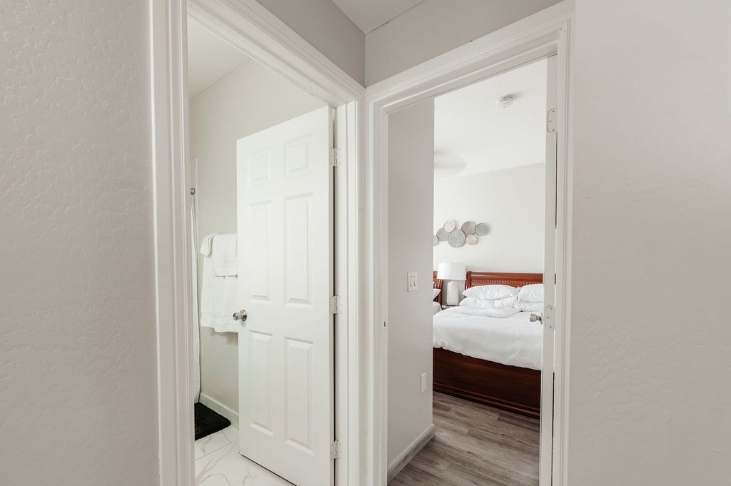 Hallway with two doorways: one to a bathroom with a closed door, the other to a bedroom with a bed.