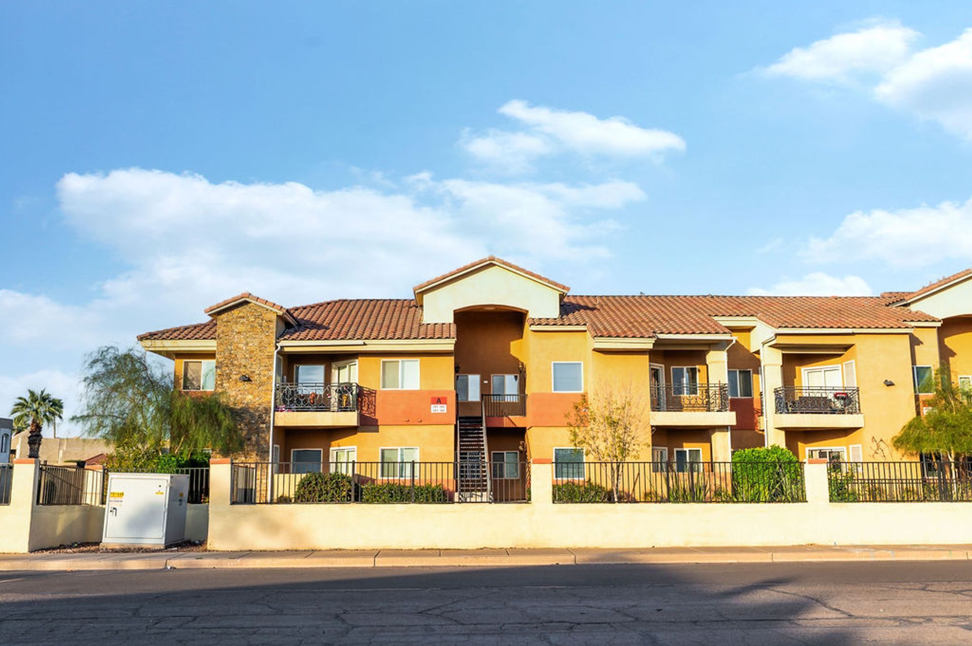Multi-story apartment building with stucco exterior, balconies, and tile roof against a blue sky.