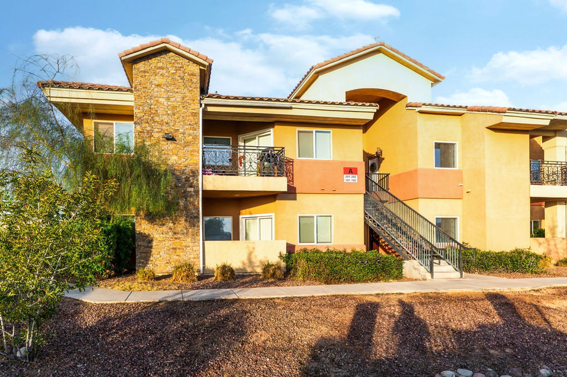 Multi-story apartment building with tan stucco exterior, stone accent, balconies, and exterior stairs under blue sky.