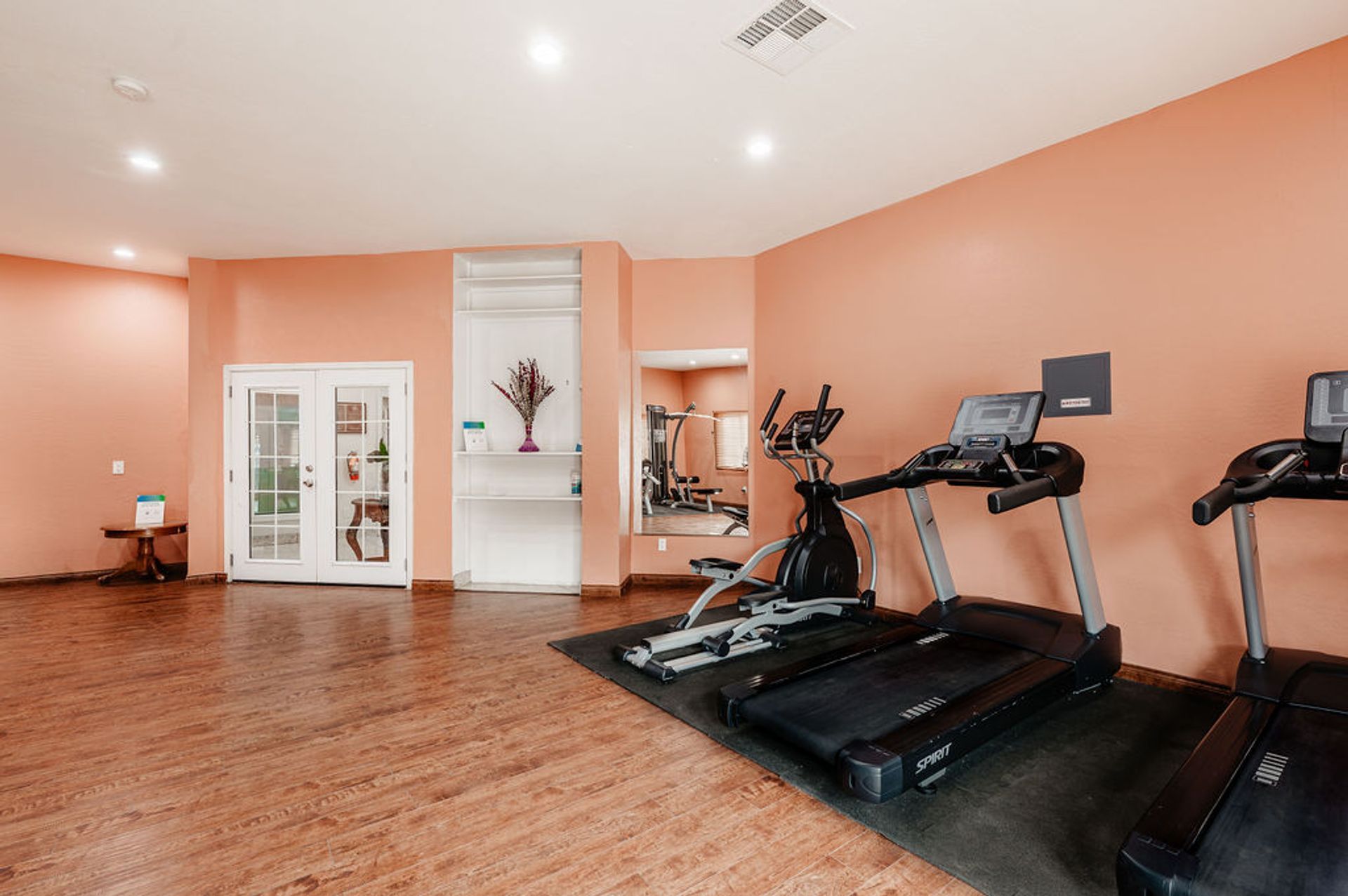 A gym with treadmills, elliptical, and weights in a room with peach-colored walls and wood flooring.