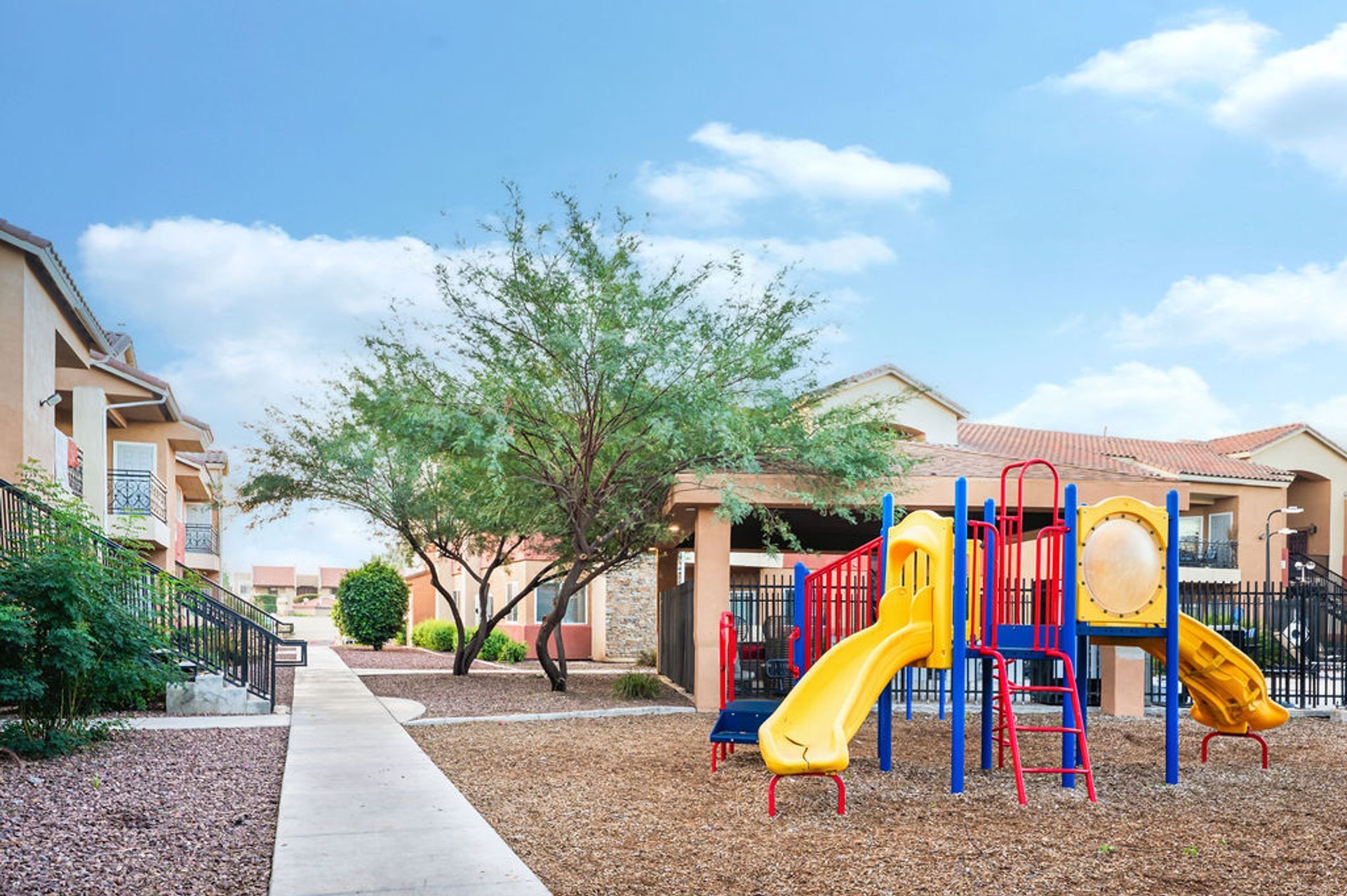 Playground with slides, surrounded by wood chips, near apartment buildings under a blue sky.