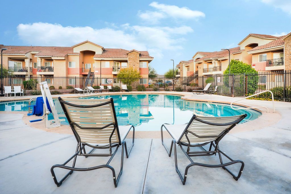 Two lounge chairs face a swimming pool, apartment buildings in background. Blue sky.