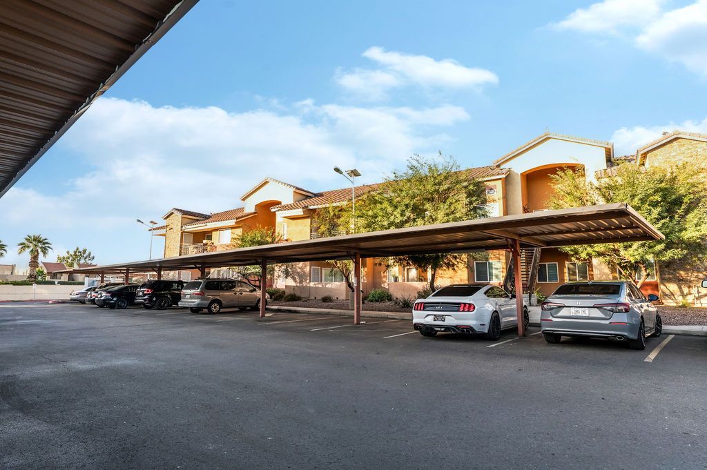 Parking area with covered carports and parked vehicles in front of apartment buildings under a blue sky.