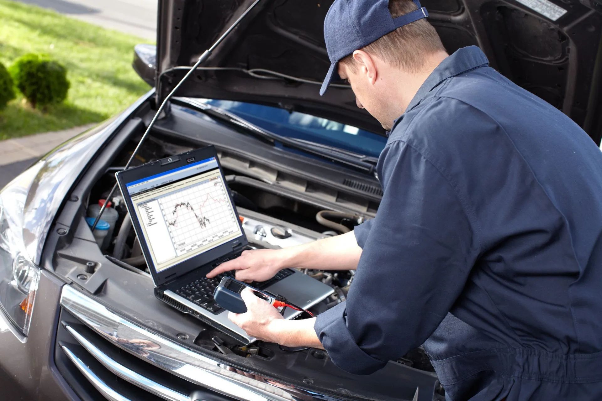 A man is working on a car with the hood open and using a laptop.