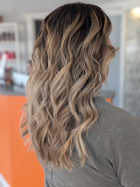 A woman with long wavy blonde hair is wearing a grey shirt who has balayage hair.