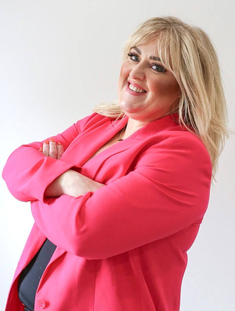 A woman in a pink jacket is smiling with her arms crossed