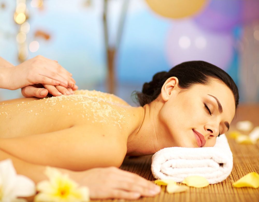 Woman receiving a back scrub at a spa. She's relaxed, eyes closed. Yellow and white flowers and a rolled towel are nearby.