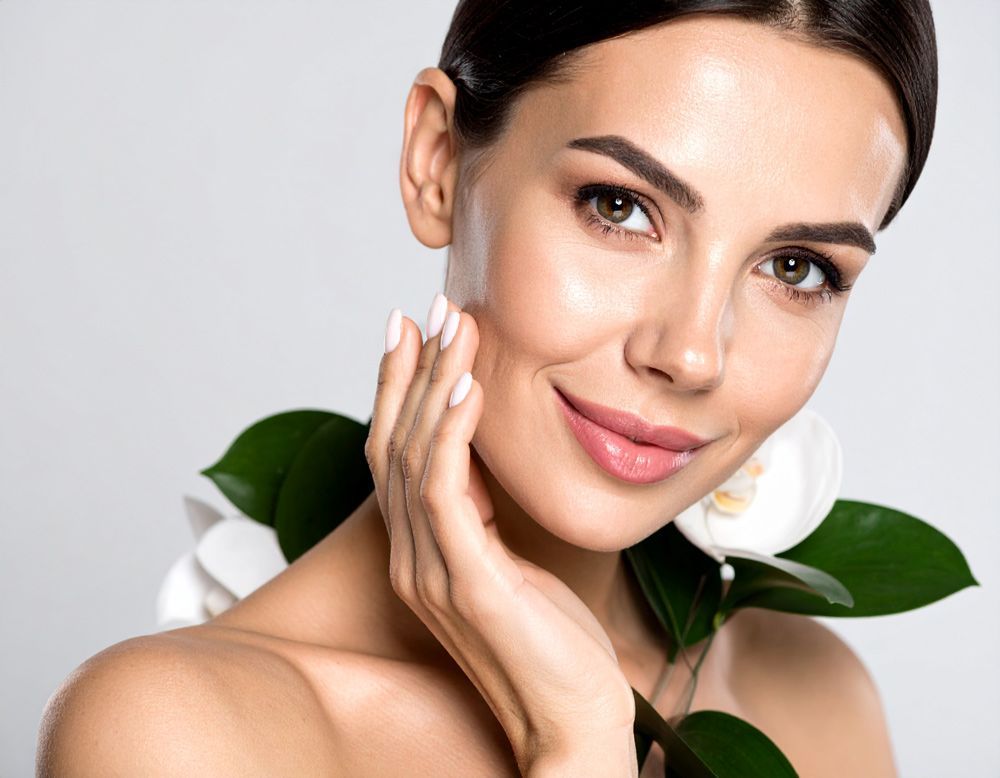 A woman is touching her glowing, shiny, clean, face.