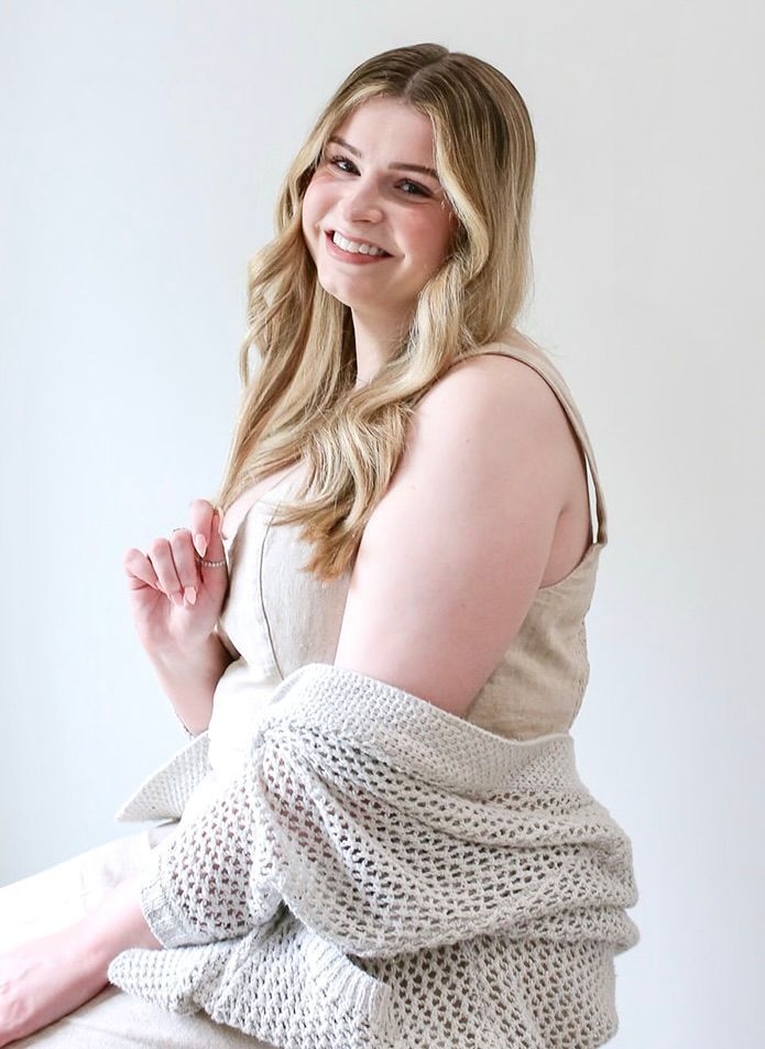 A woman is sitting on a stool wearing a sweater and smiling.