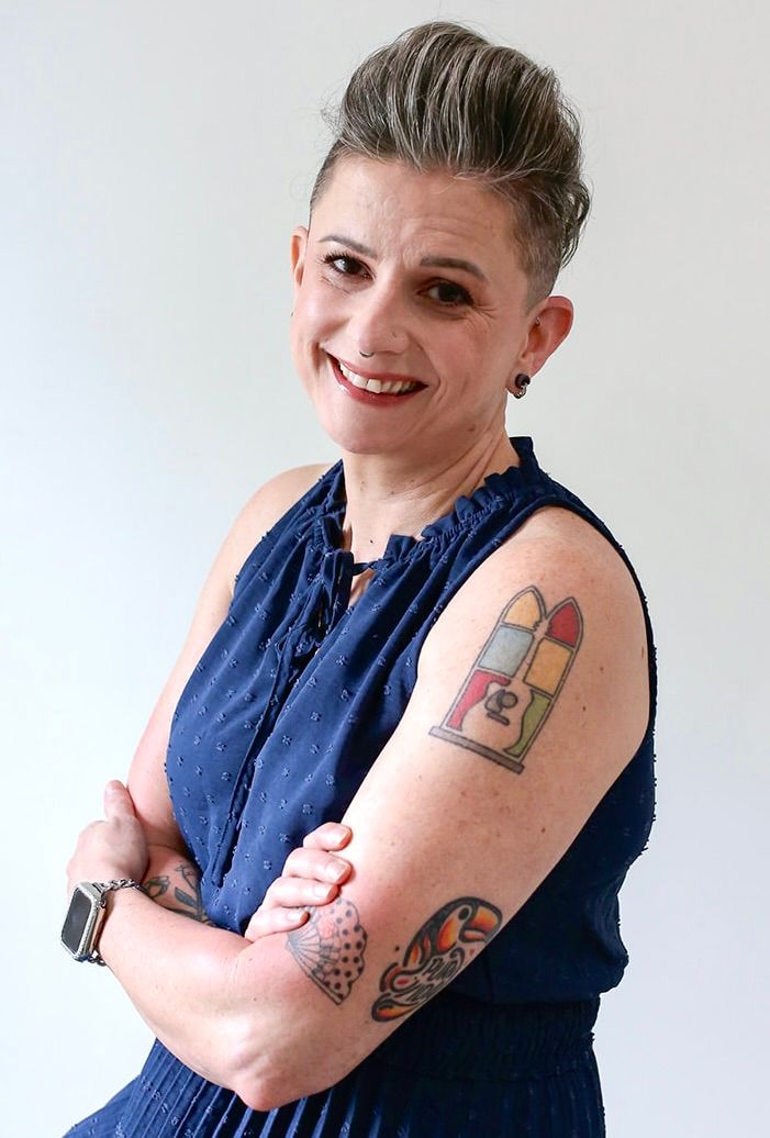A woman with tattoos on her arm is smiling and standing with her arms crossed.