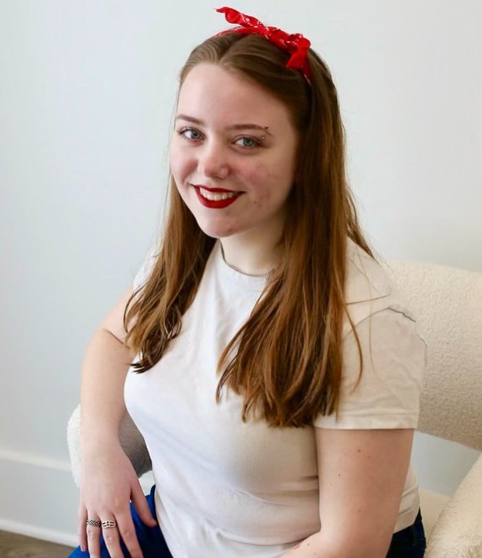 Woman with red lipstick, bandana, in white shirt, smiles, sits on chair.