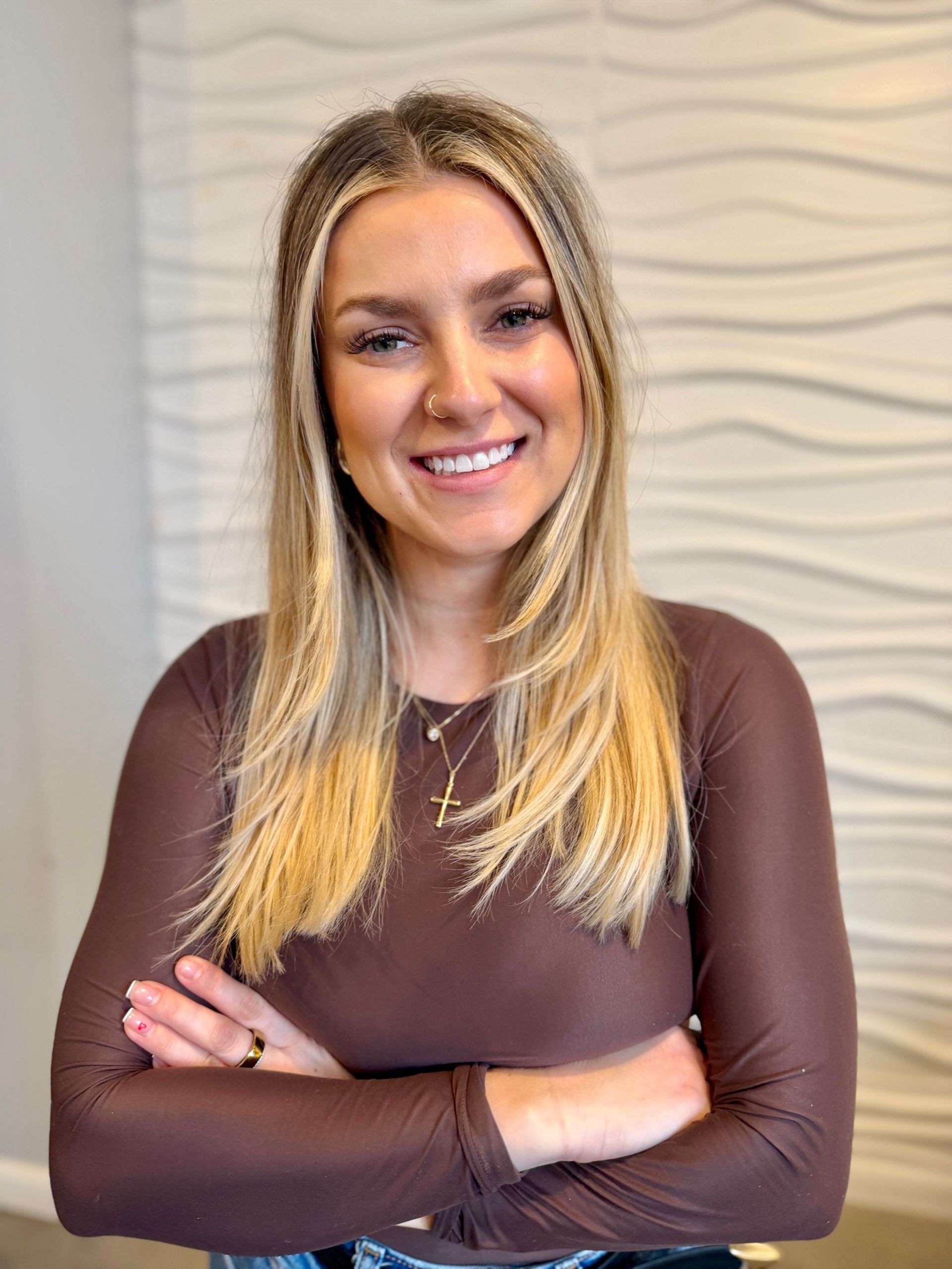 Woman with blonde hair, smiling, wearing a dark blue one-shoulder top, arms crossed. Sitting in an off-white chair.