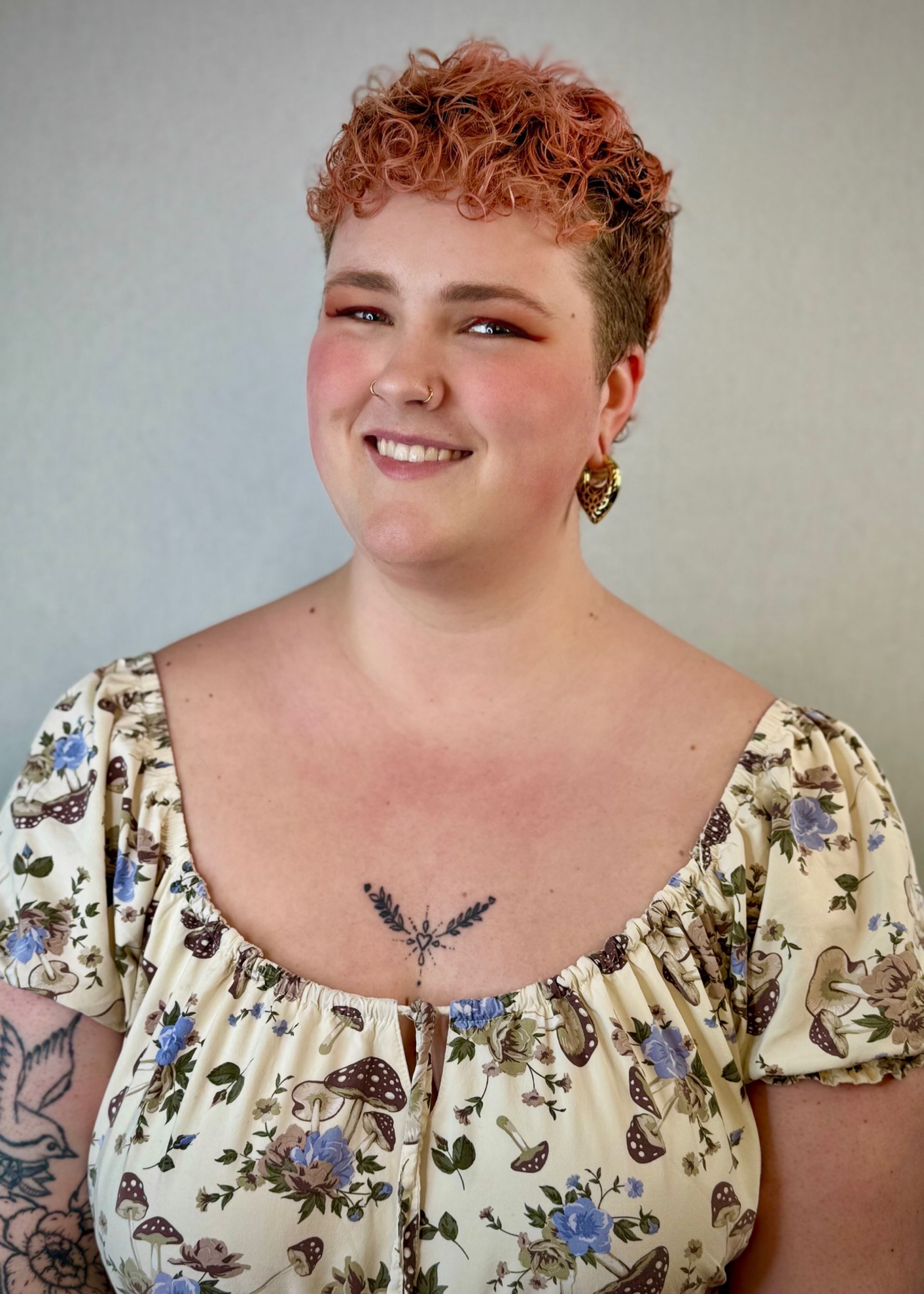 Woman with short pink hair smiles. Wearing floral top, heart earrings, and chest tattoo.