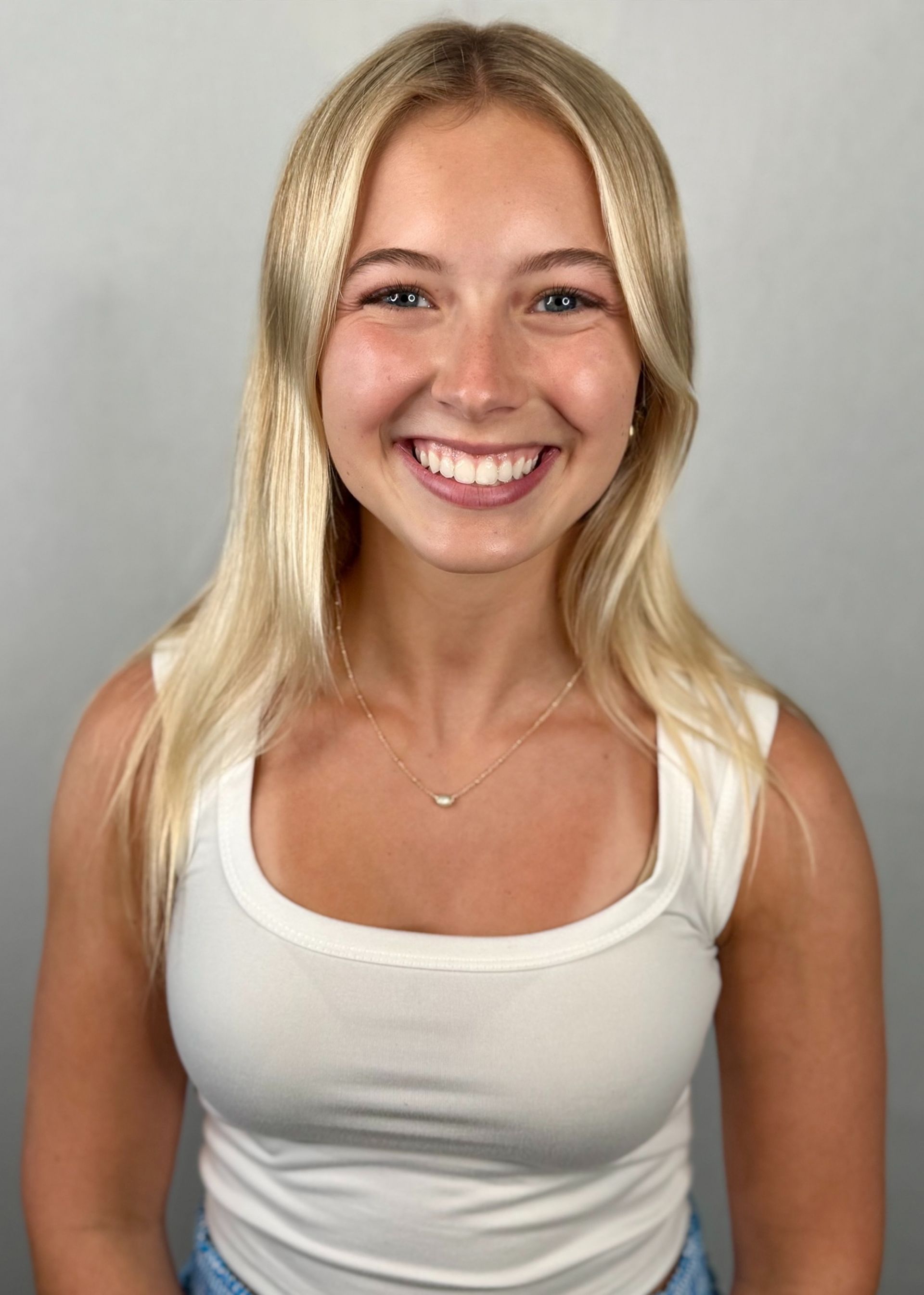 Blonde woman with a wide smile wearing a white tank top. Light gray background.