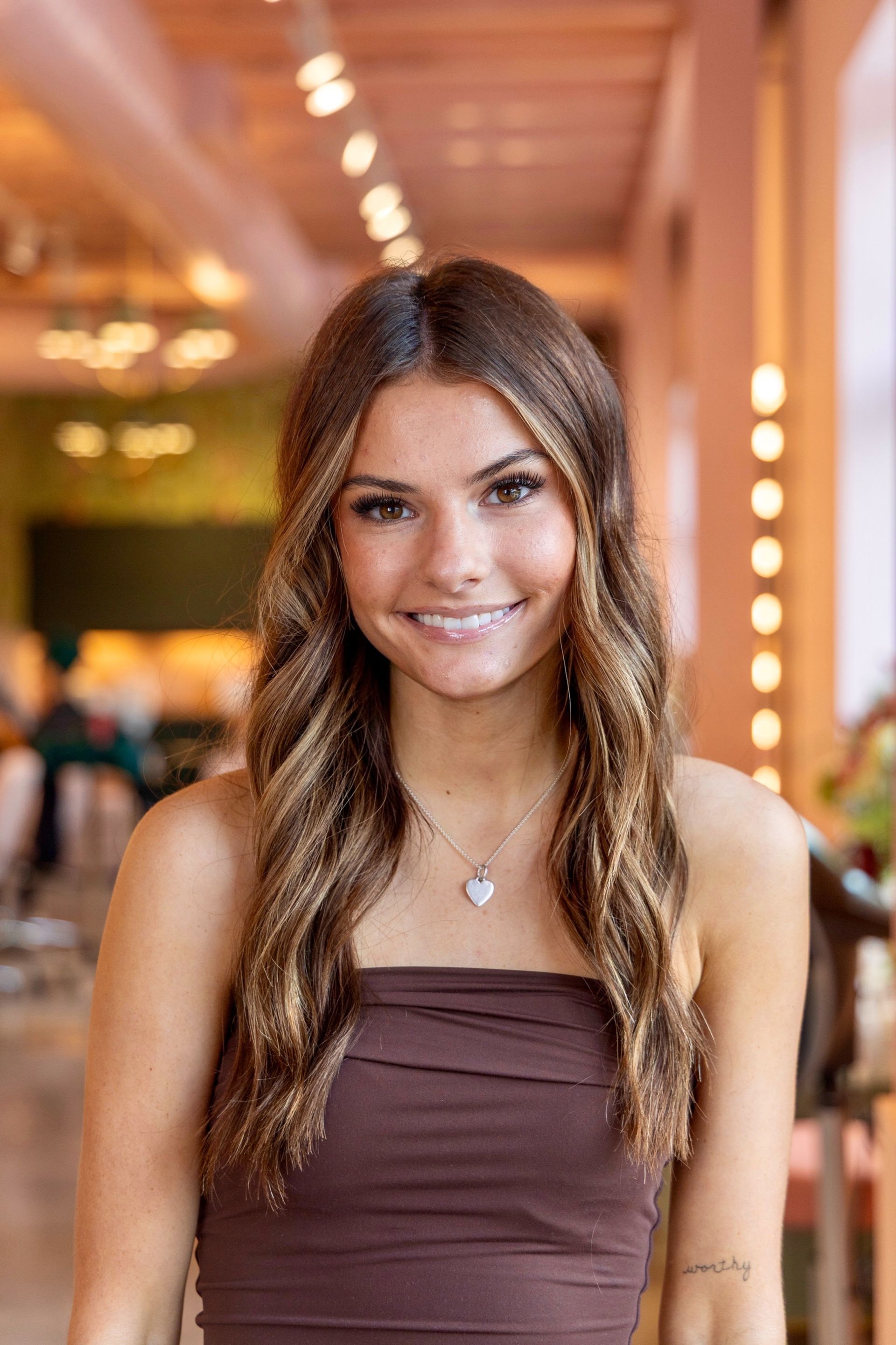 Woman with long brown hair in a pink top smiles with hand on hip. White background.