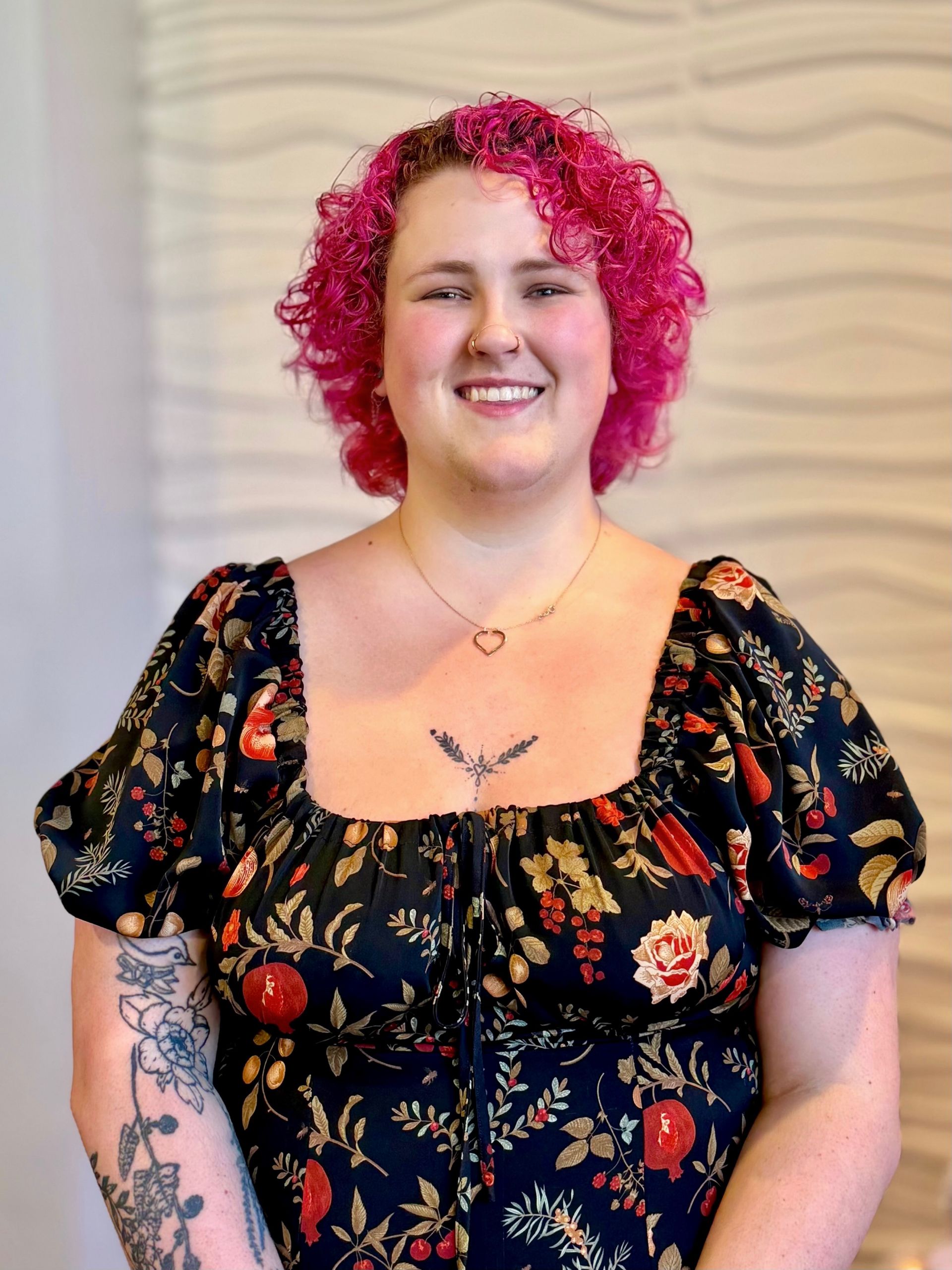 Woman with short pink hair smiles. Wearing floral top, heart earrings, and chest tattoo.