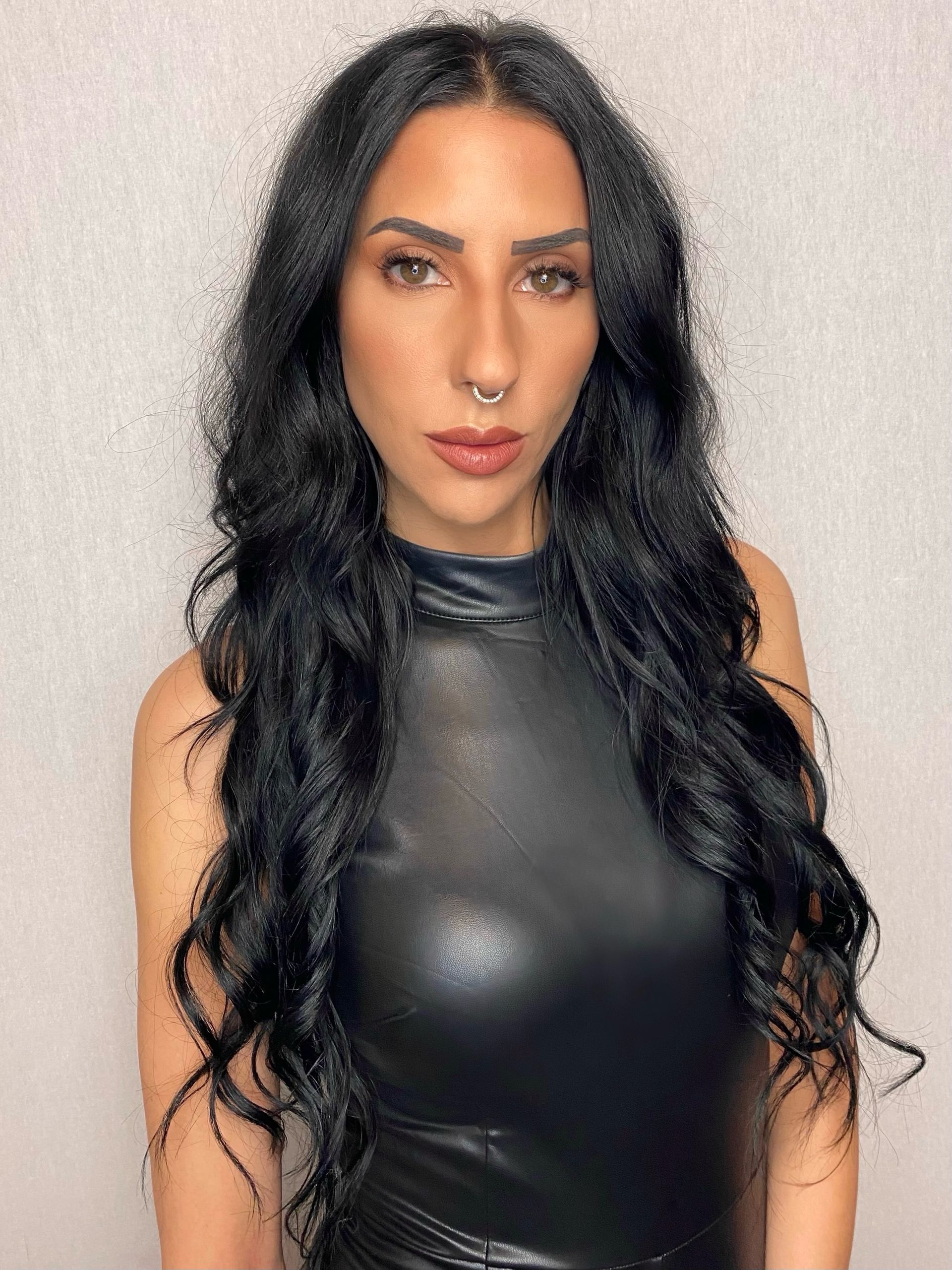 A woman with long black hair and a nose ring is wearing a black leather dress.