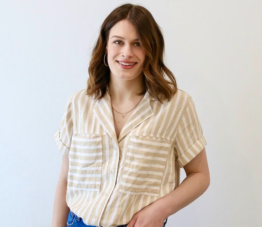 A woman wearing a striped shirt and jeans is standing in front of a white wall.