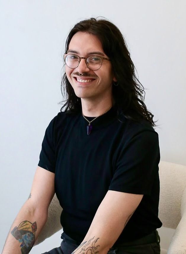 A man with long hair and glasses is sitting in a chair and smiling.