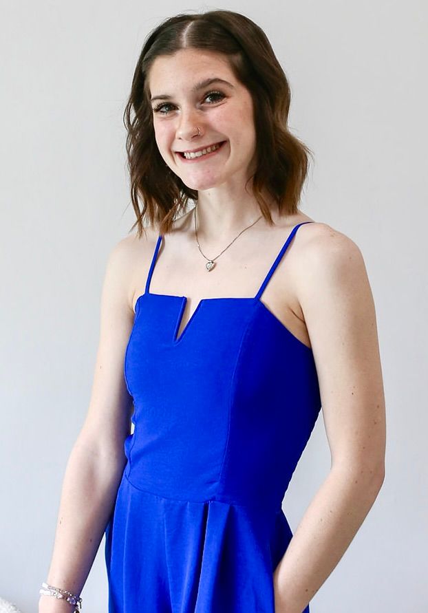 A woman in a blue dress is smiling and standing with her hands in her pockets.
