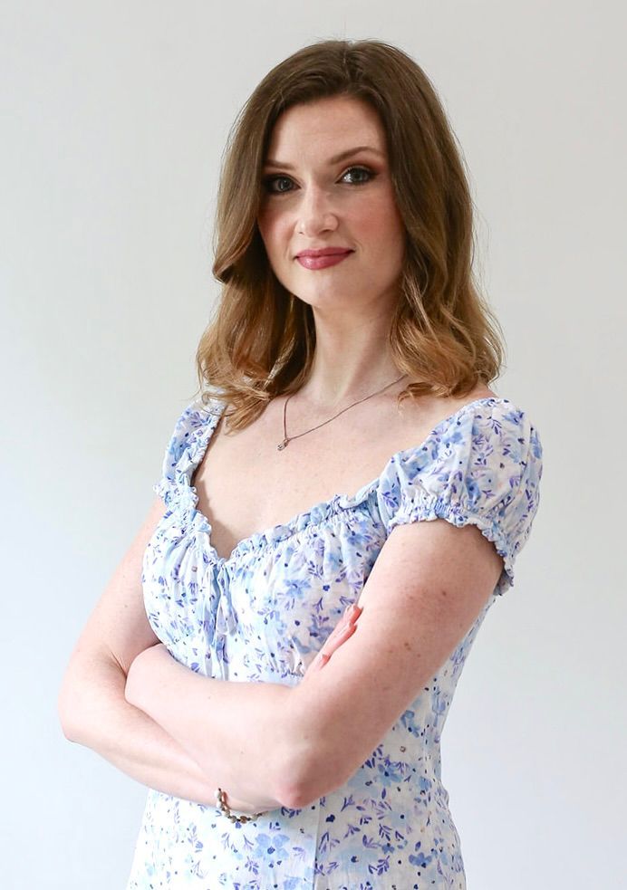 A woman in a blue and white floral dress is standing with her arms crossed.