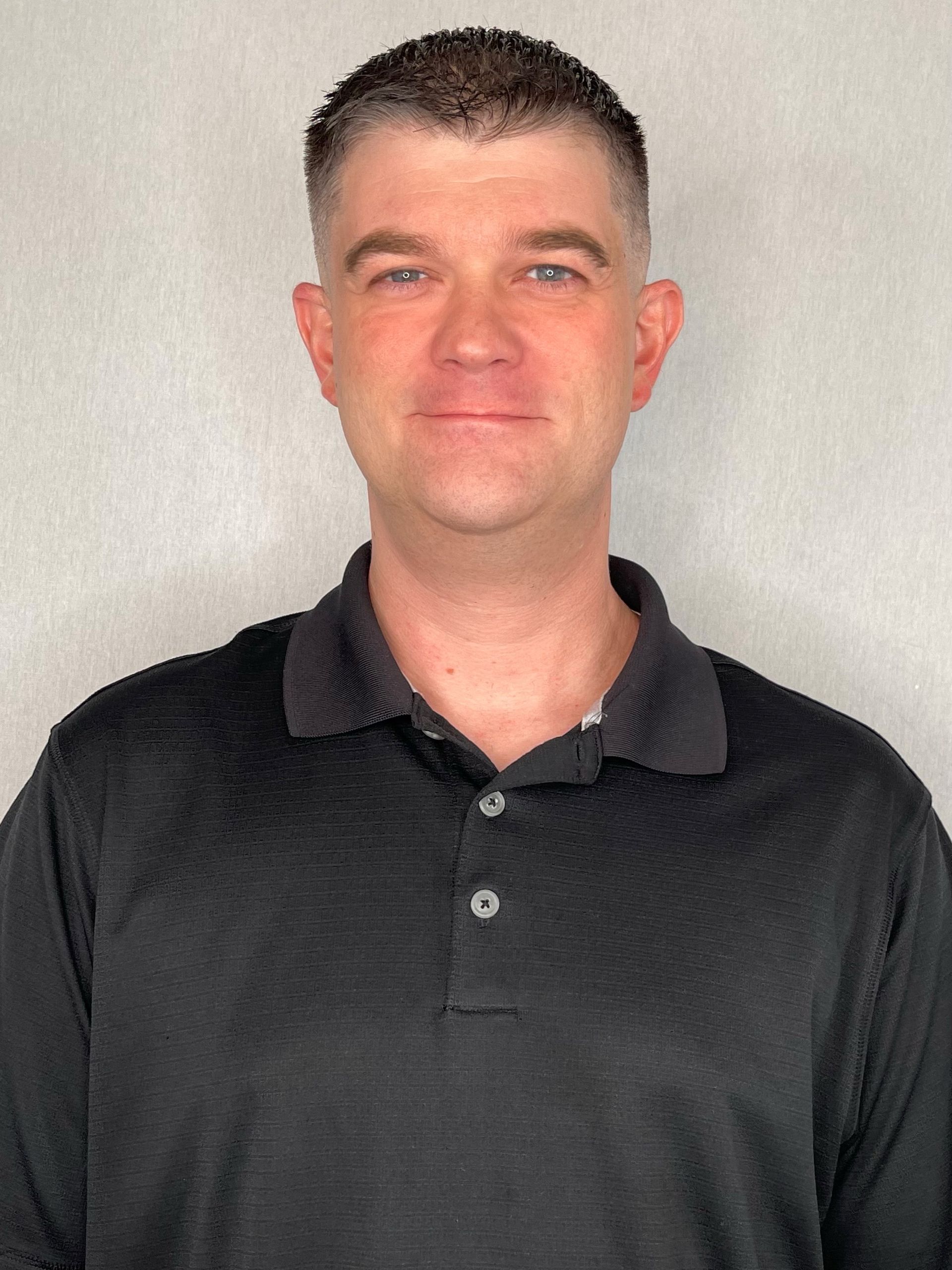 A man wearing a black polo shirt is standing in front of a white wall.