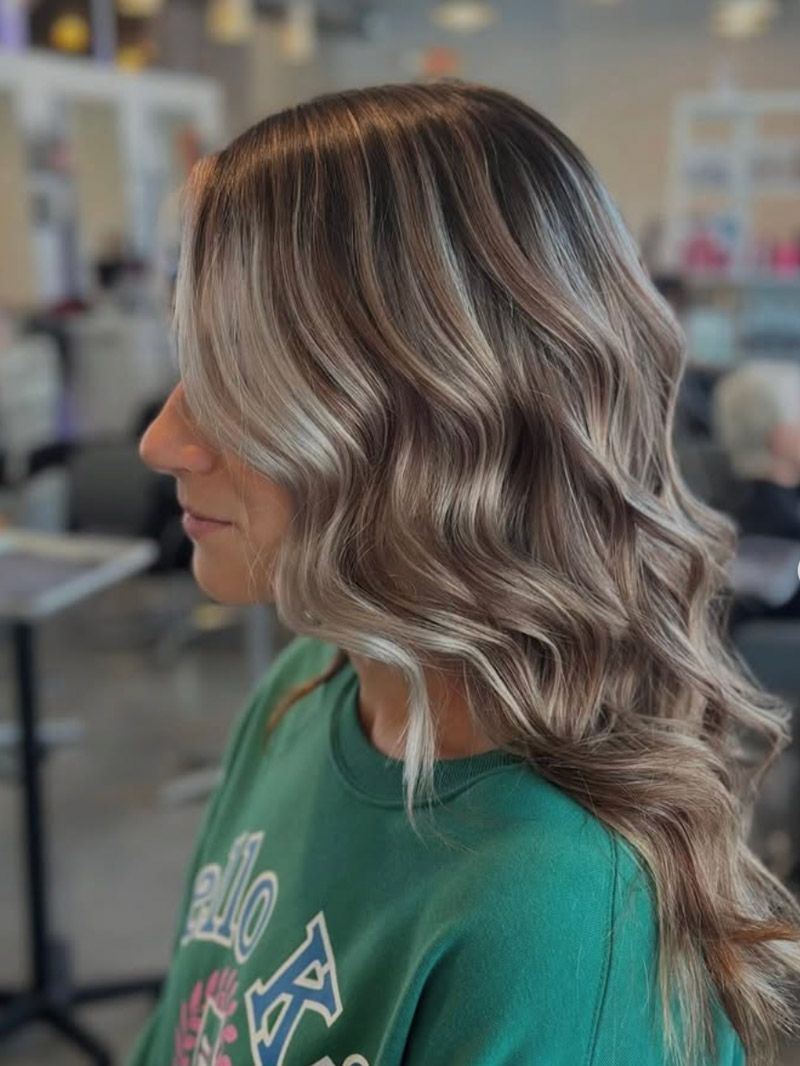 Side profile of a person with long, wavy, ash-brown and blonde highlighted hair at Luxe Salon and Spa.