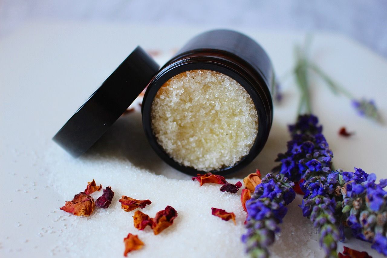 Open jar with sugar scrub, lavender, and rose petals on a white surface.