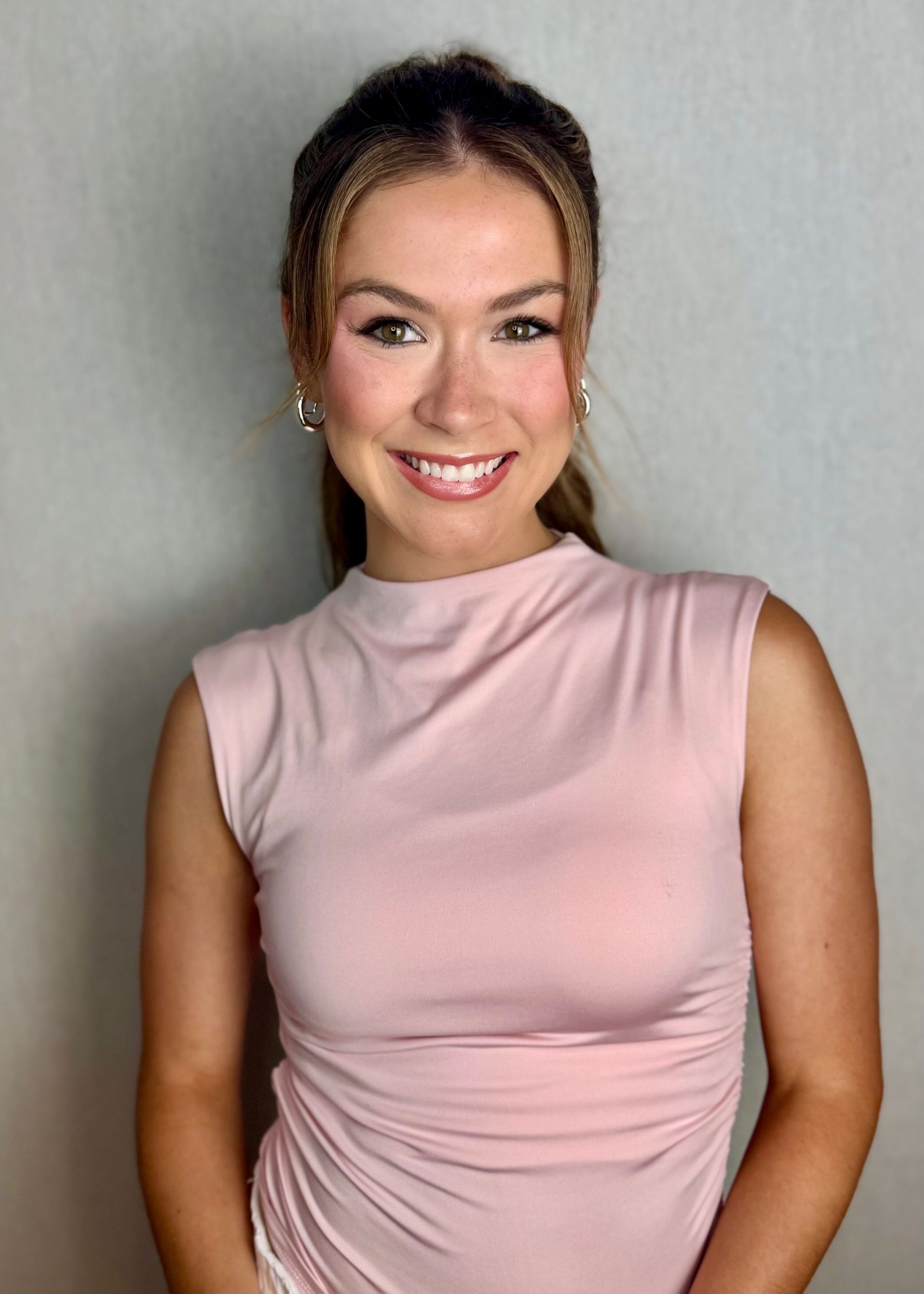 Woman smiles at the camera, wearing a pink sleeveless top; neutral background.
