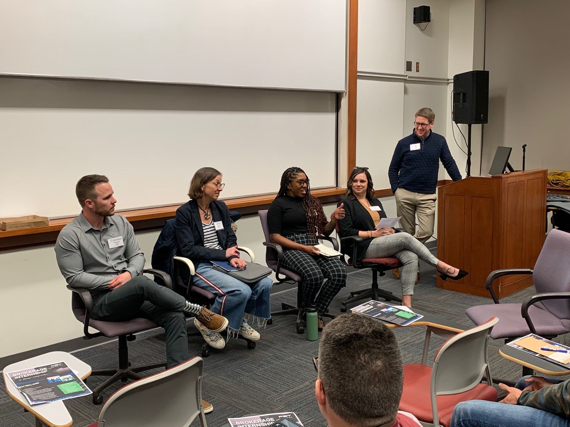 A group of people are sitting in chairs in front of a podium in a classroom.