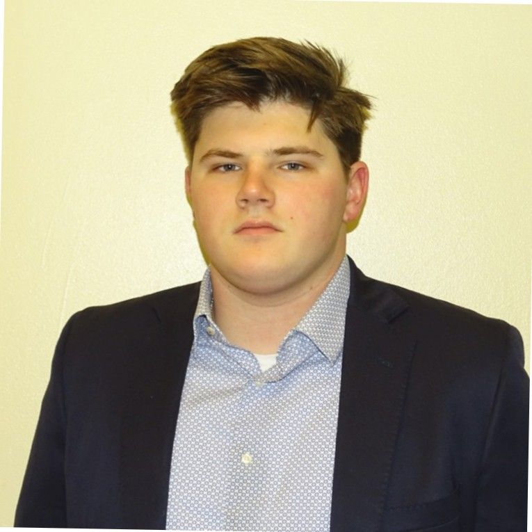 A young man in a suit and shirt is standing in front of a white wall.