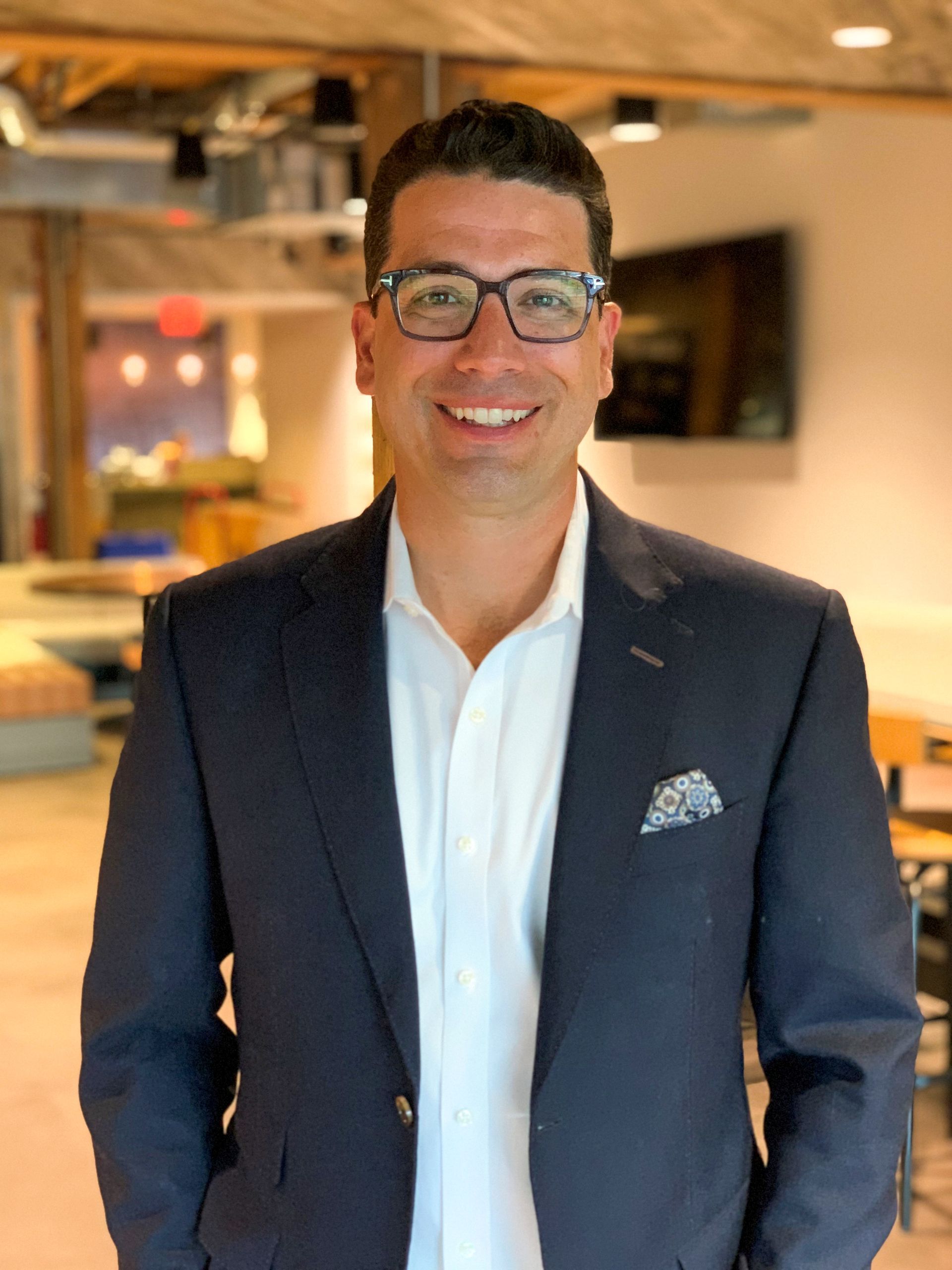 A man in a suit and glasses is standing in a room with his hands in his pockets.