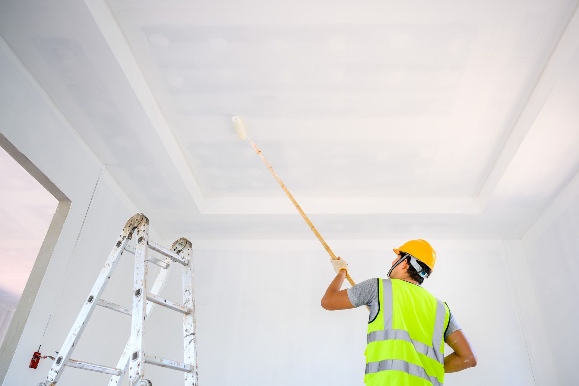 A man is standing on a ladder painting the ceiling of a room.