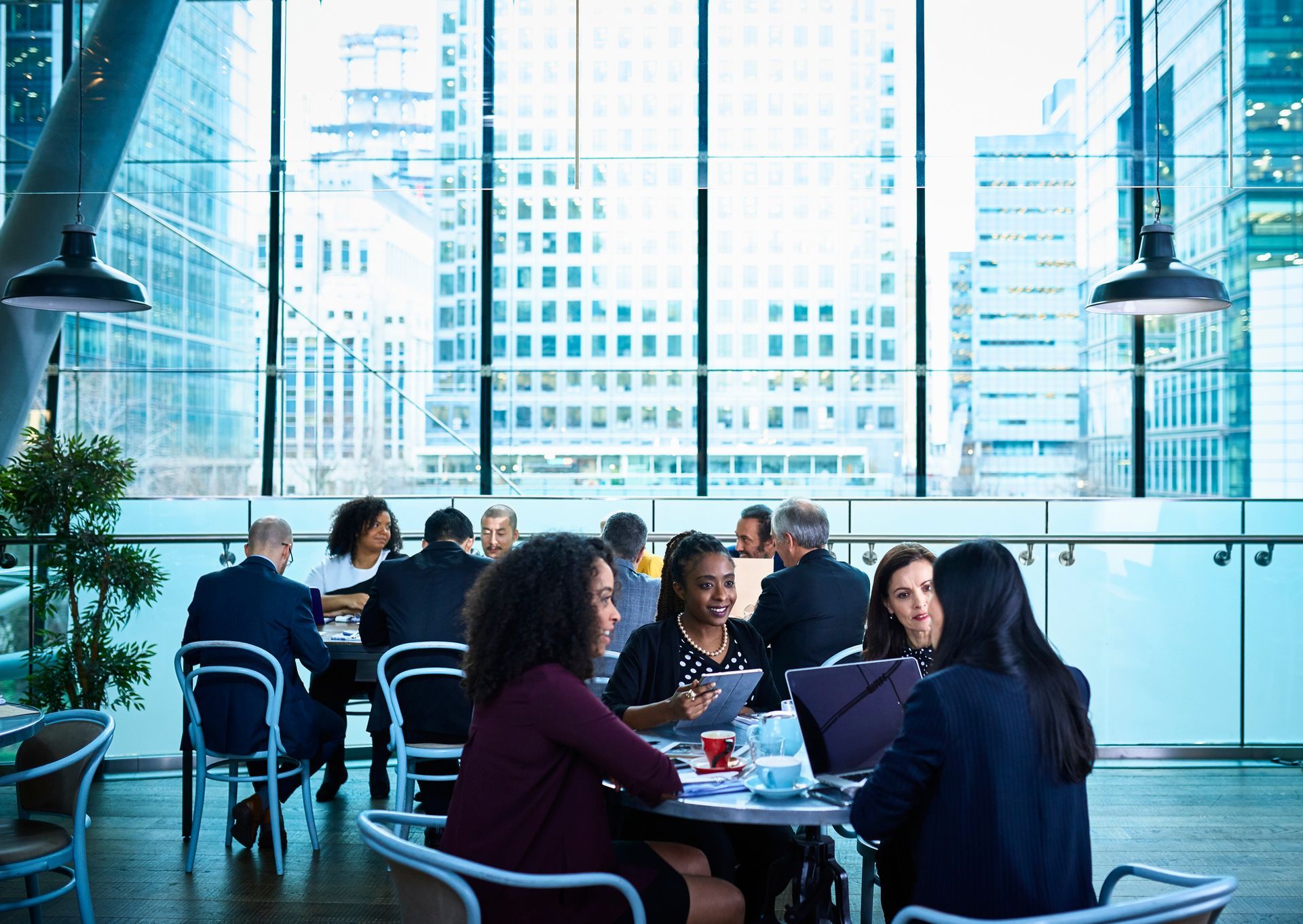 People in suits sit around tables in a modern cafe with a city view.