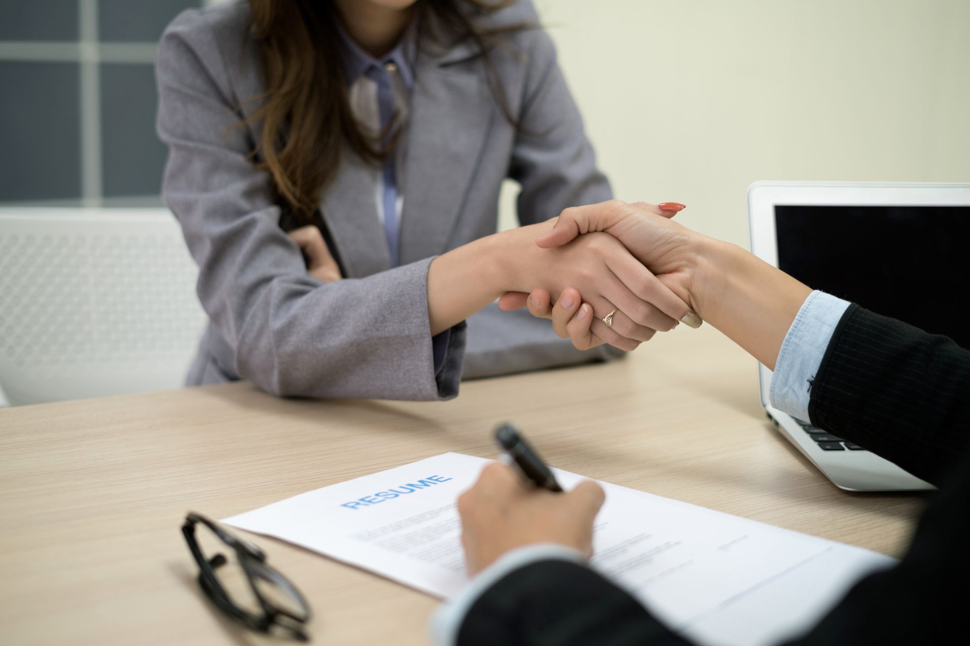 Two women shaking hands over a contract on a desk.