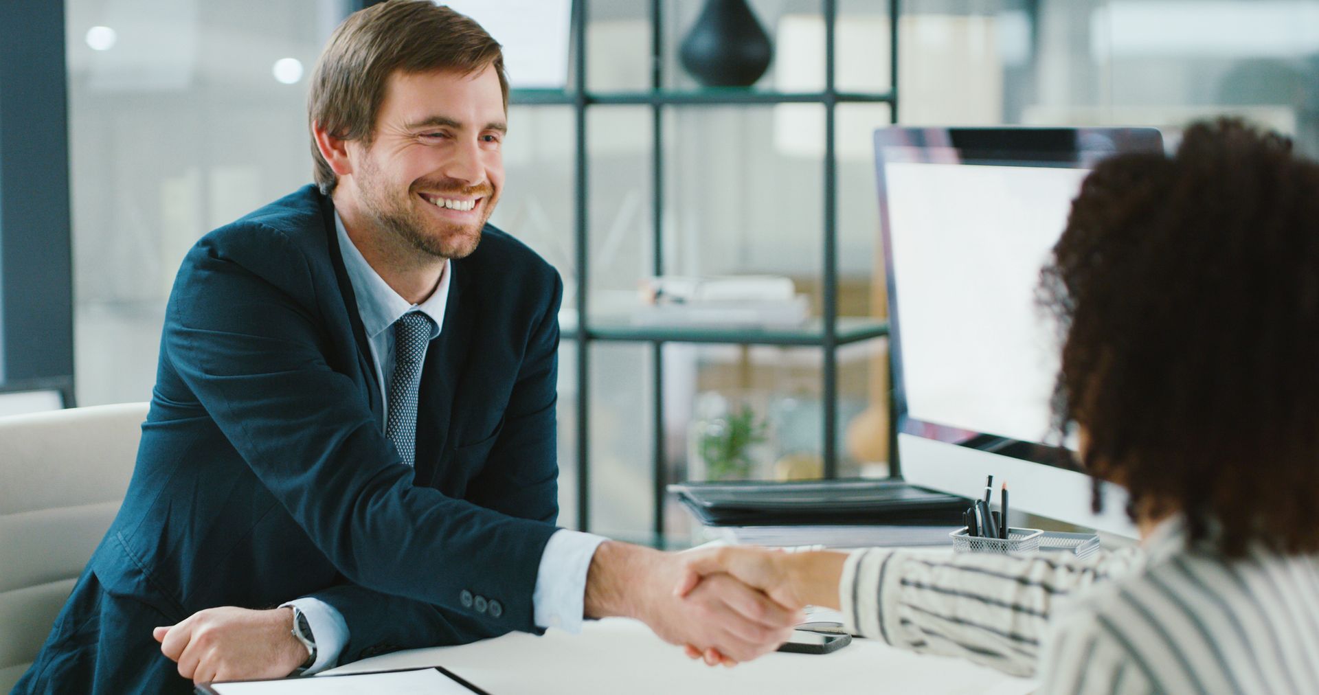 Man in suit shakes hands with a woman at an office desk; both smiling.