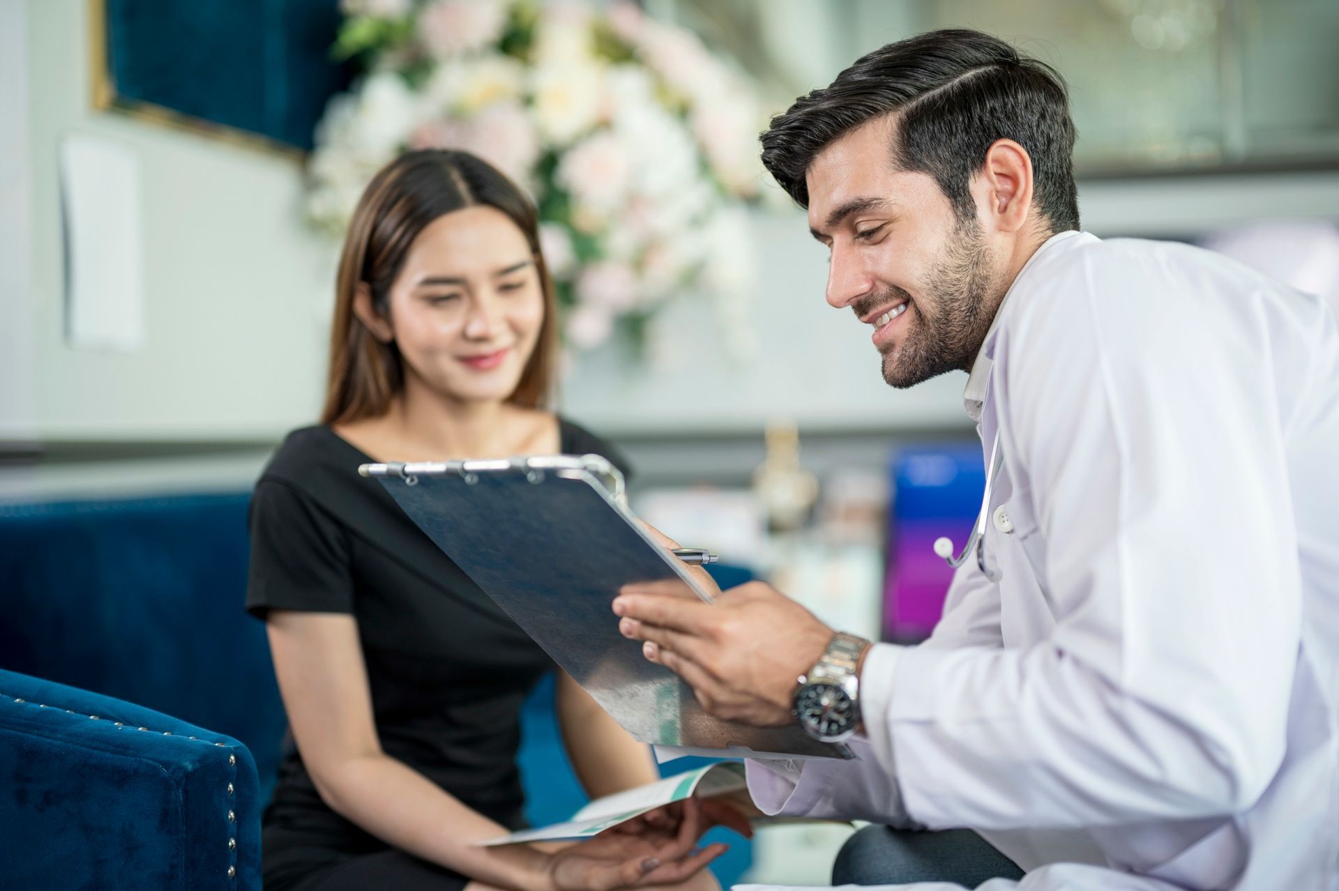 A doctor in a white coat reviews a clipboard with a smiling female patient in a clinic setting.