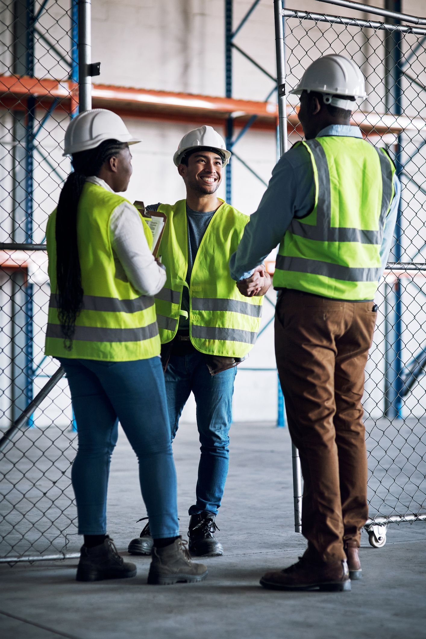 Three warehouse workers in vests and hard hats, talking in a storage area.