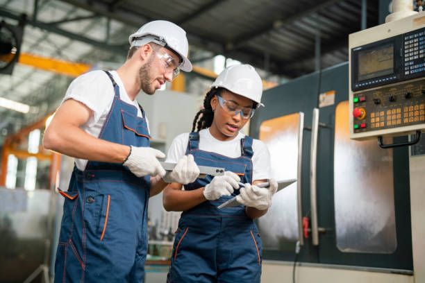Two factory workers wearing safety gear inspect a metal part next to a CNC machine.
