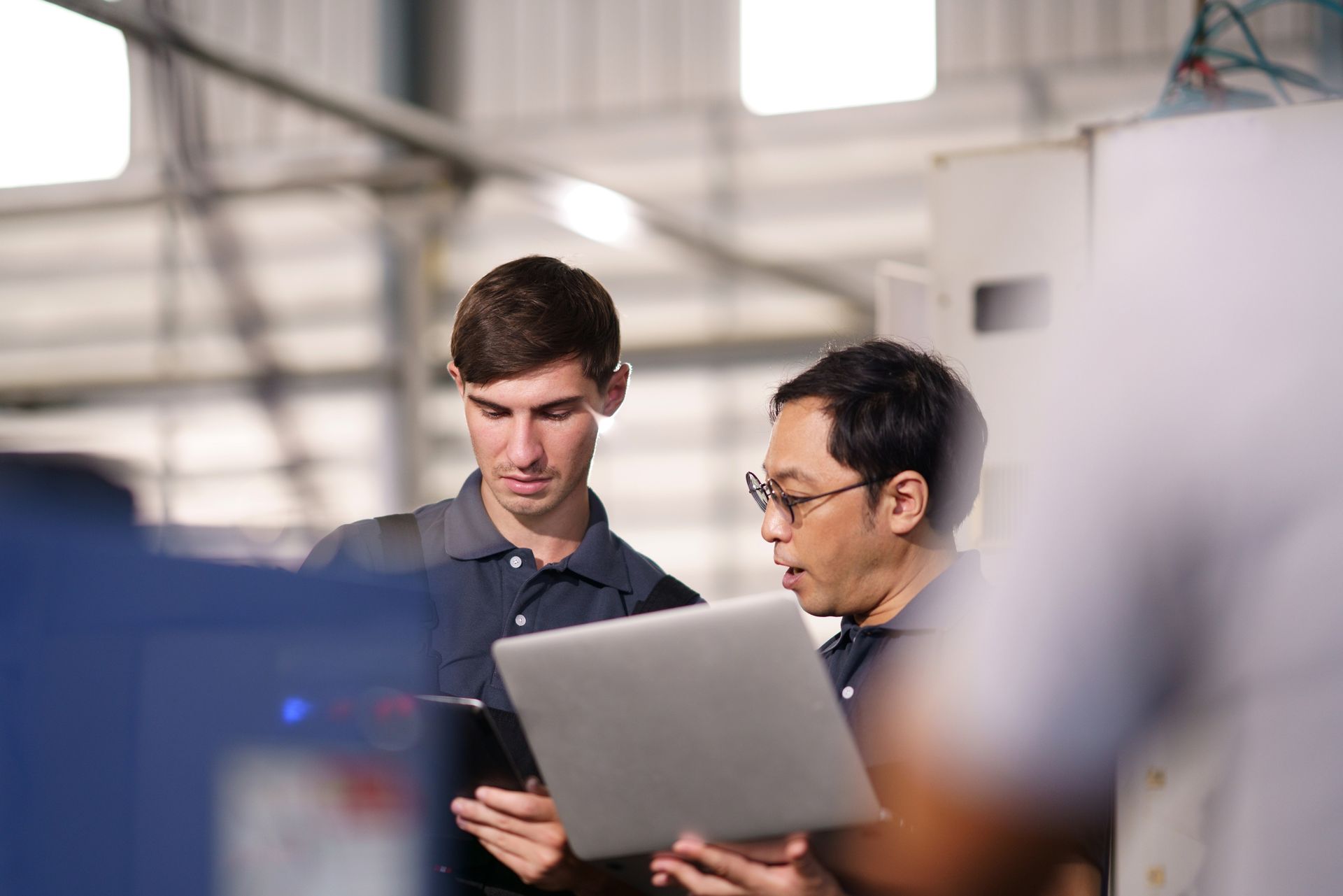 Two men, one young and one Asian, examining a laptop and tablet in a factory setting.