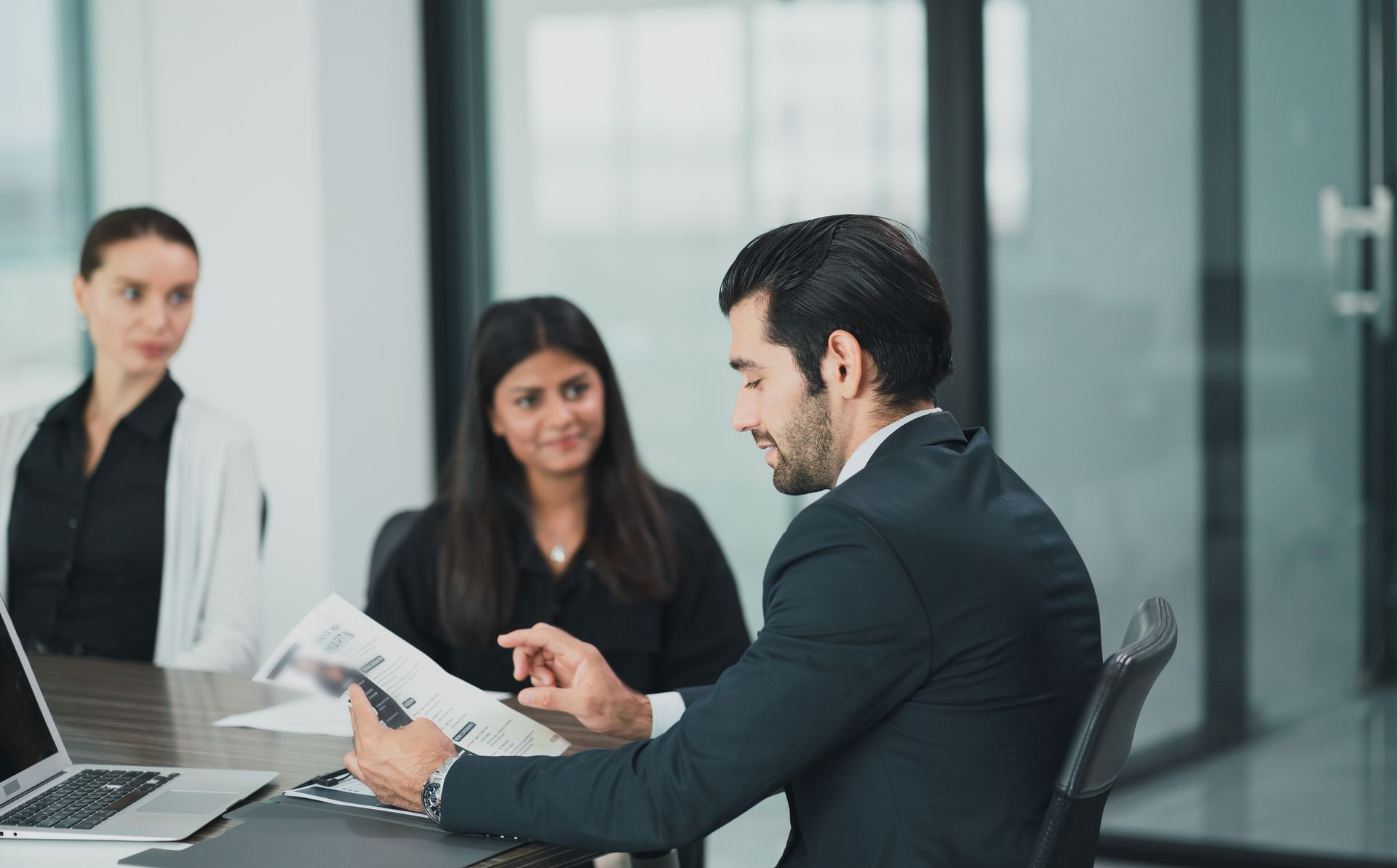 Three business people in a modern office, looking at documents and laptop; man pointing.