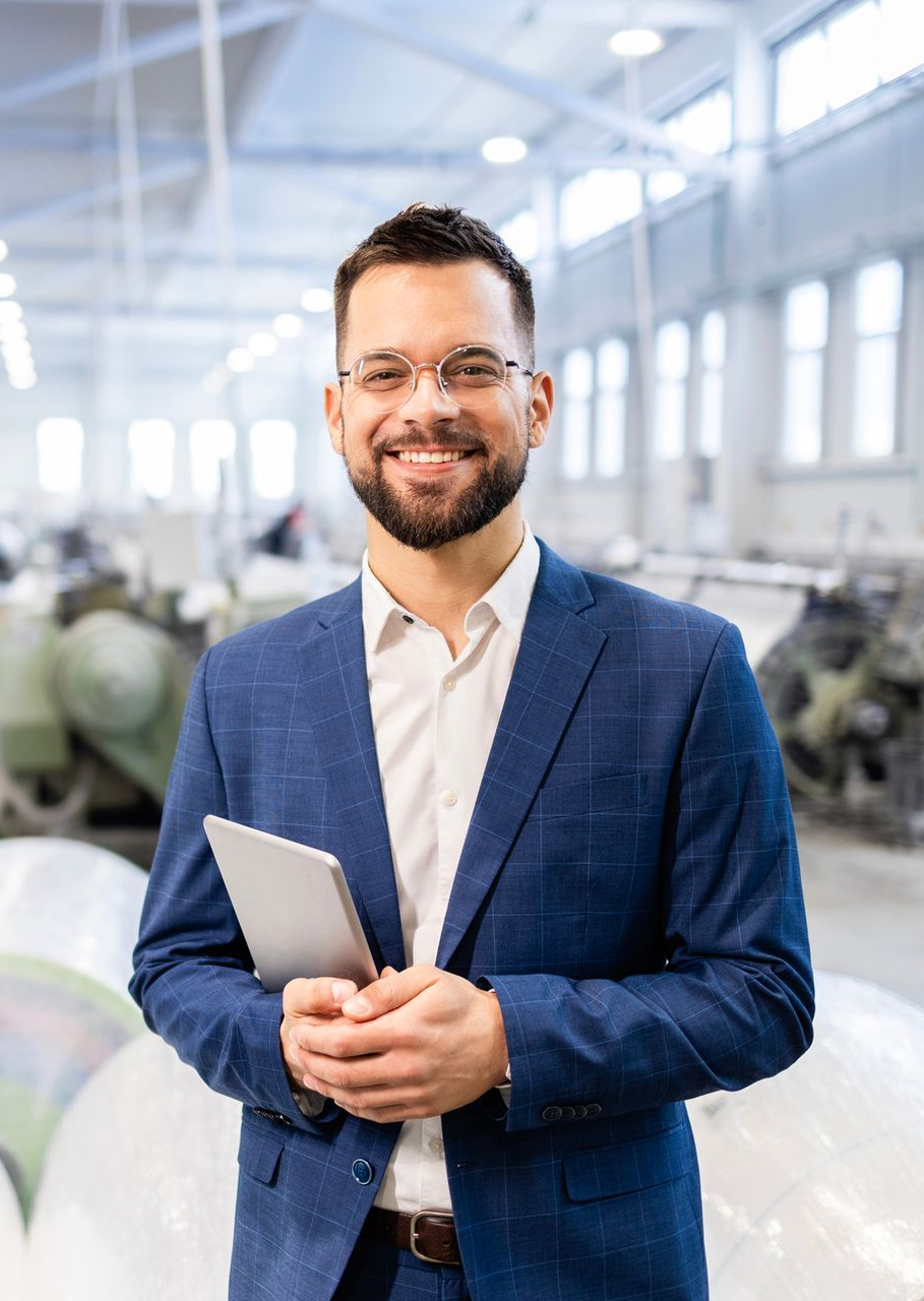 Man in a blue suit smiles, holding a tablet in a factory setting.
