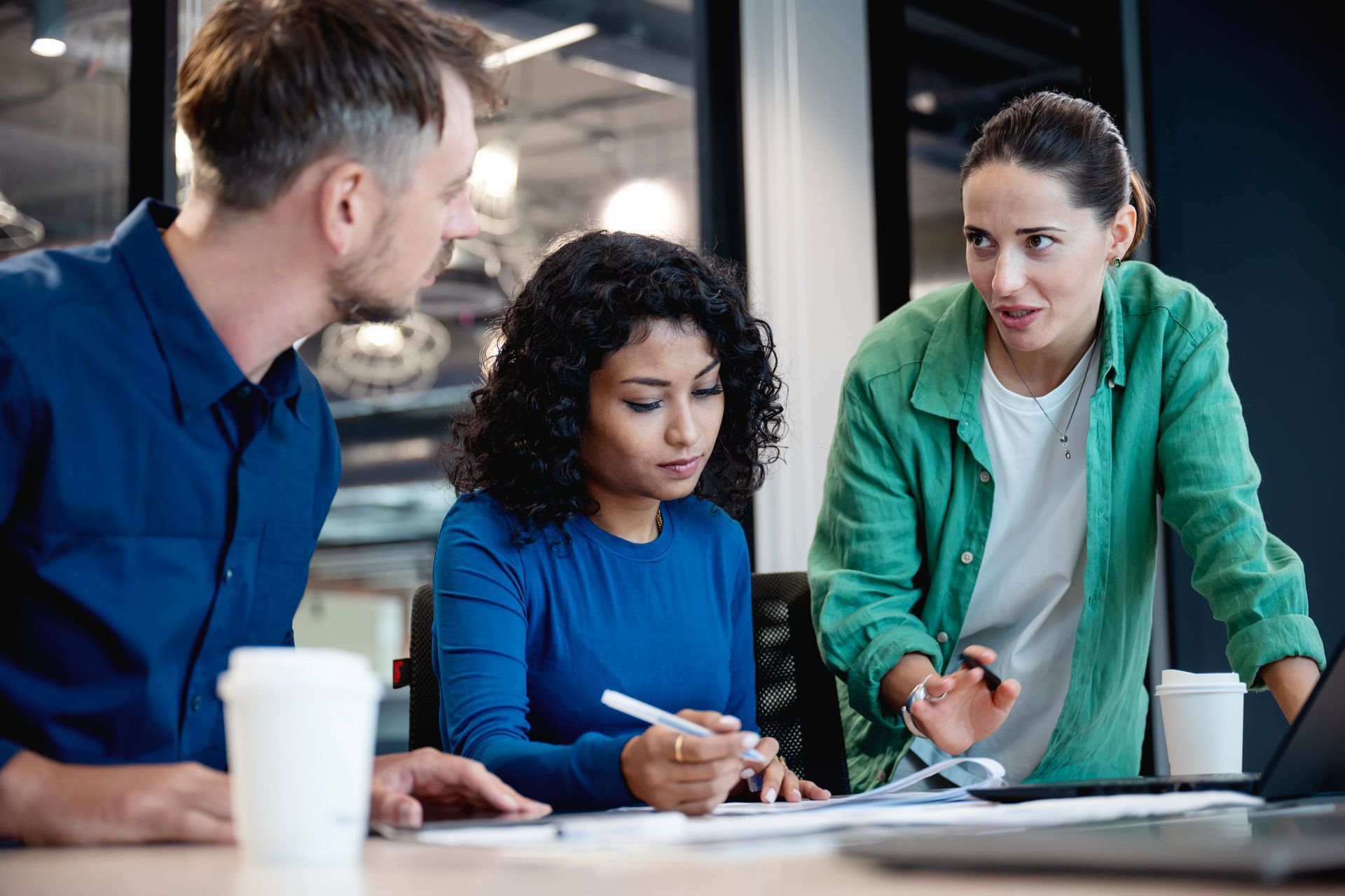 Three people collaborating over documents in an office setting. Woman gestures, two others focused.