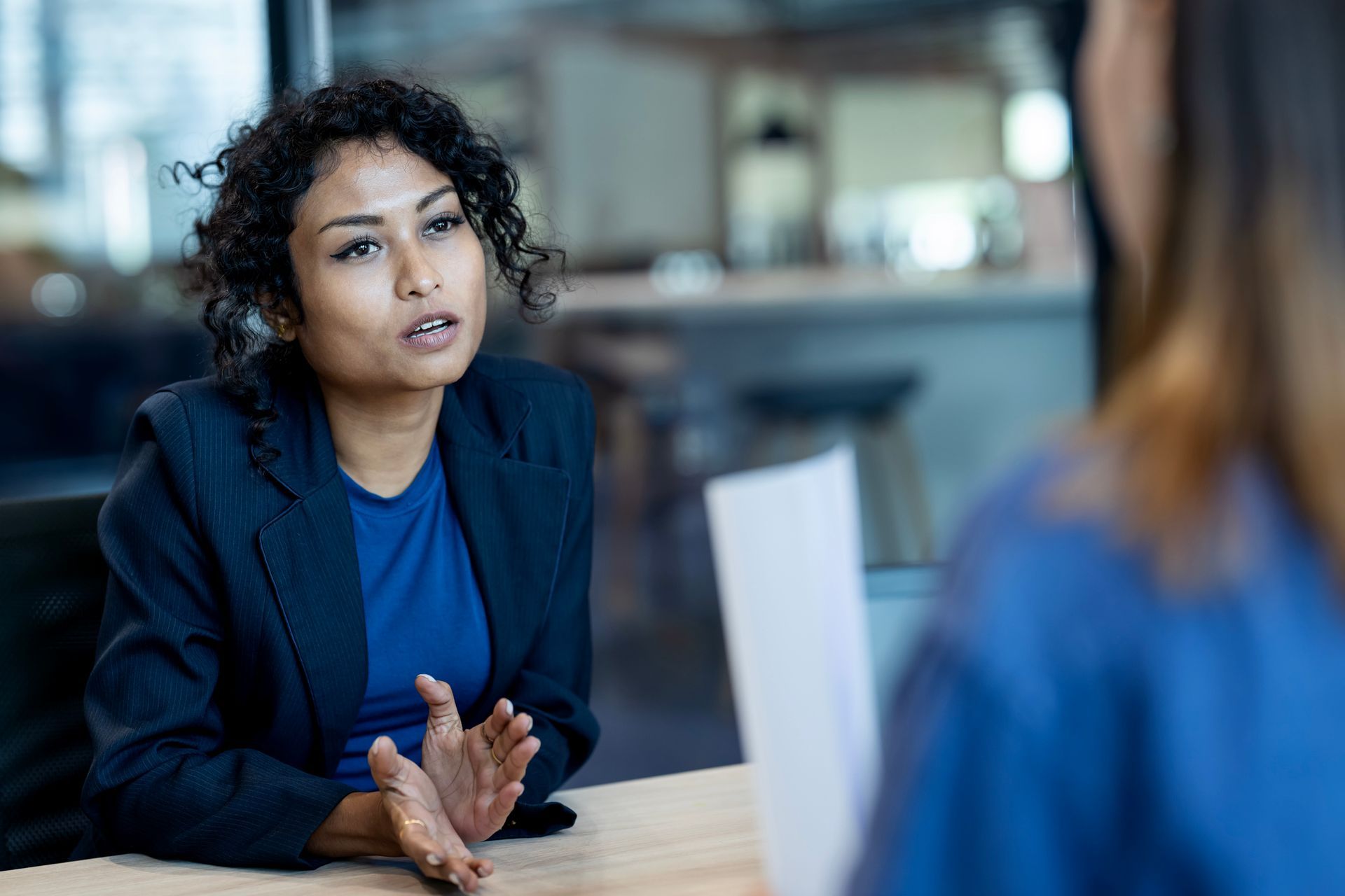 Woman in a blue blazer gestures while speaking during an interview, across a table from another person.