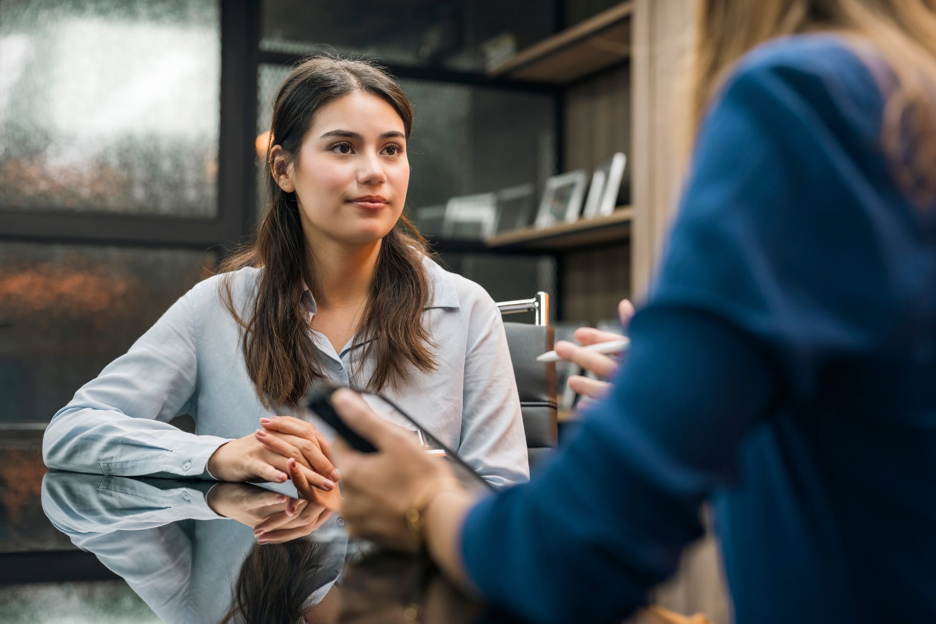 Woman at a table in an office setting, facing another person, appears to be listening during an interview.