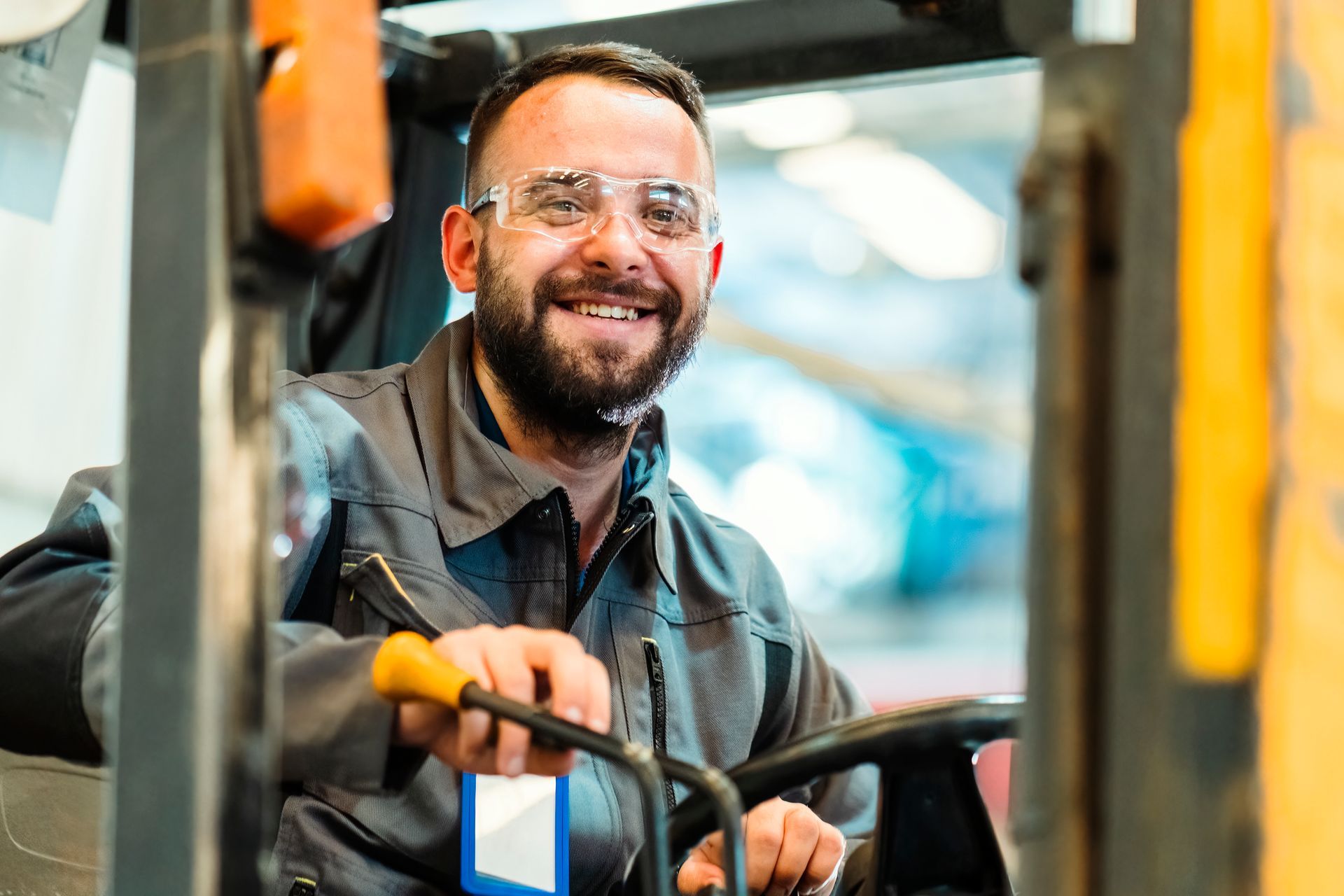 Man in safety glasses smiles while driving a forklift in a warehouse.